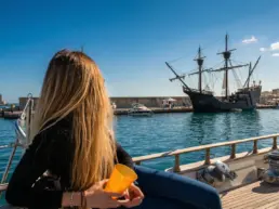 Blonde woman seen from behind with a glass watching a historic wooden galleon moored in the harbour