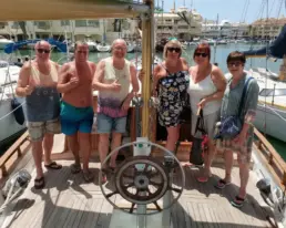 Seis adultos posando con el pulgar arriba junto al timón de un velero en la dársena de Benalmádena