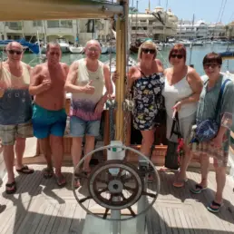 Six adults posing with thumbs up by a yacht's helm in the Benalmádena marina
