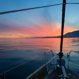 Atardecer de cielo rosa y violeta reflejado en un mar en calma visto desde la proa de un velero