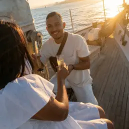Pareja joven de blanco brindando con copas de cava a contraluz dorado en la cubierta de un velero