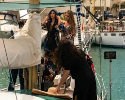 Dos modelos con vestidos elegantes y una estilista durante un rodaje de moda en un velero en puerto