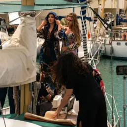 Dos modelos con vestidos elegantes y una estilista durante un rodaje de moda en un velero en puerto