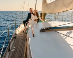 Pareja de novios abrazada sobre la borda de teca de un velero en mar abierto al atardecer