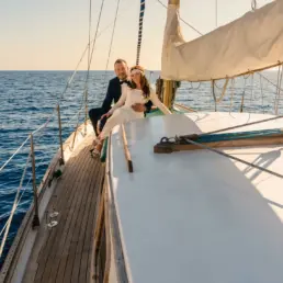Pareja de novios abrazada sobre la borda de teca de un velero en mar abierto al atardecer