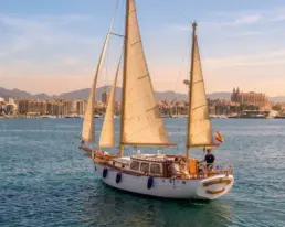 Velero navegando a vela a la hora dorada frente a una ciudad costera con una gran catedral