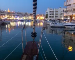 Dársena de Benalmádena al anochecer con las luces del puerto reflejadas en el agua, desde la proa