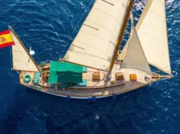 Overhead aerial view of a yacht sailing under full sail over a deep blue sea with a Spanish flag