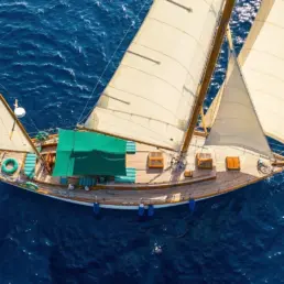 Overhead aerial view of a yacht sailing under full sail over a deep blue sea with a Spanish flag